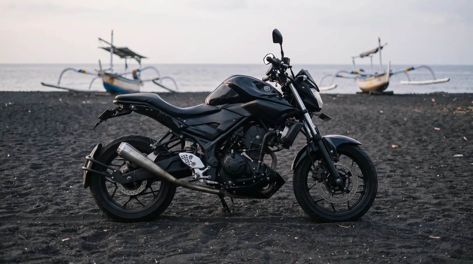 Yamaha MT-25 scrambler on a black-sand beach with traditional jukung boats, Bali