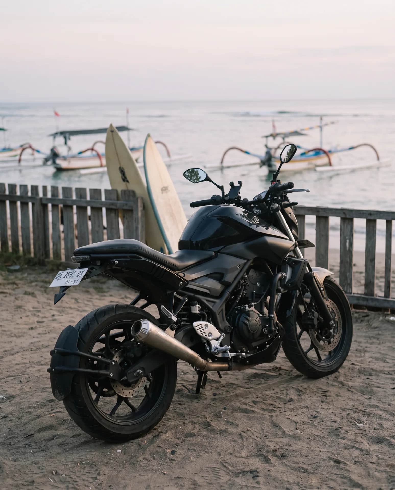 Custom scrambler Yamaha MT-25 by surfboards and outrigger boats on a Bali beach at dusk