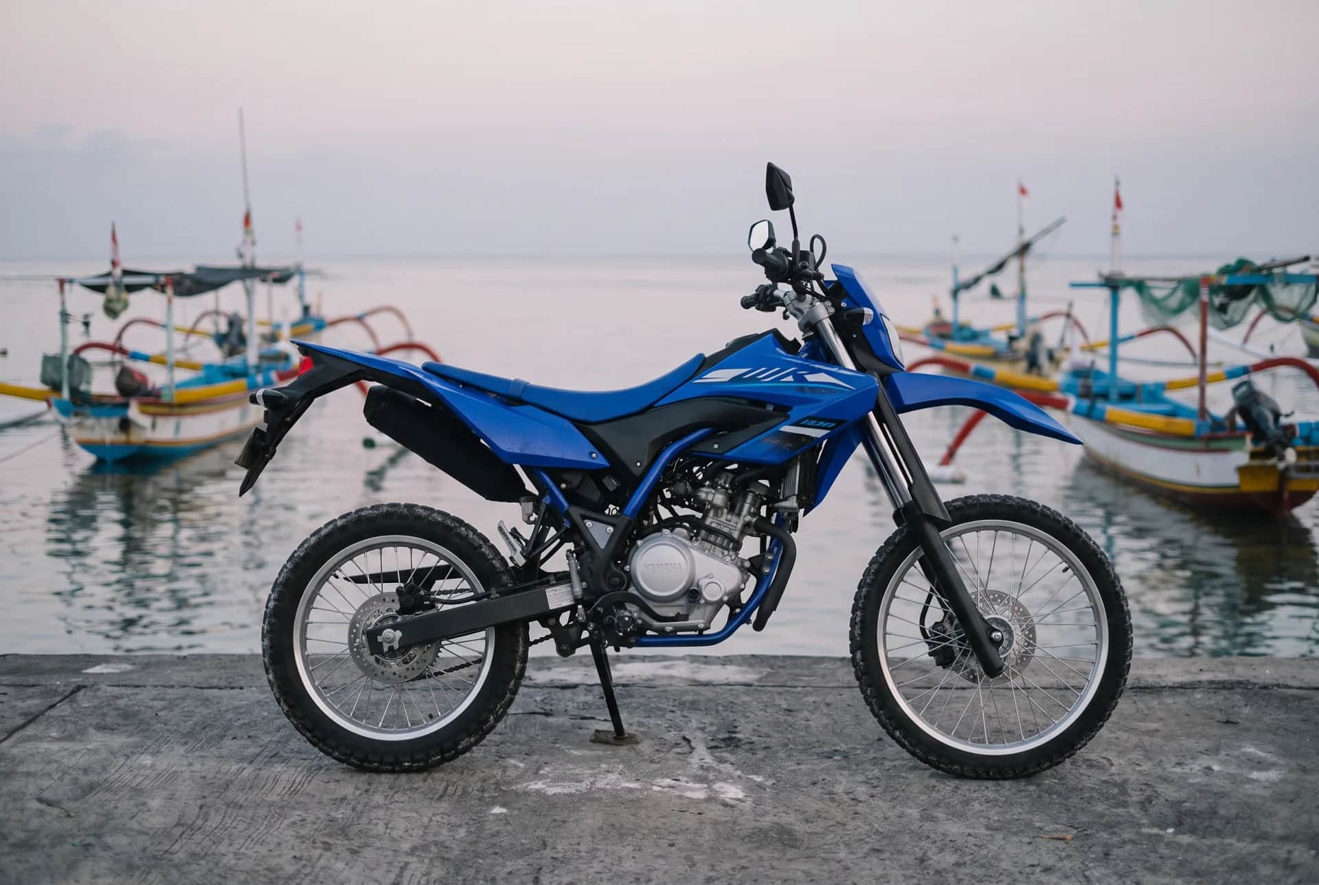 Blue Yamaha WR155 parked on a coastal dock at dusk in Bali with colourful fishing boats on calm water